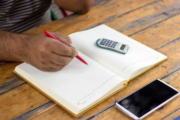 A person using a phone calculator and writing on a notepad, surrounded by papers and a laptop, illustrating the process of calculating living expenses for an emergency fund.