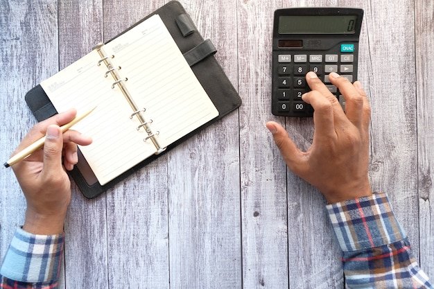 A close-up of a hand holding a calculator over a table with a budget spreadsheet, a pen, and various bills scattered around. The focus is on the calculator's display showing a calculation of income after taxes.