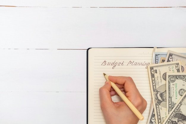 A hand writing a budget breakdown on a notepad, with various bills and financial documents scattered around the table. The focus should be on the organization and planning involved in budgeting.