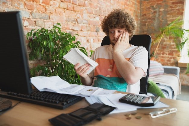 A person sitting at a desk, looking stressed while reviewing bills and financial statements on a laptop. The scene is set in a small, cluttered apartment, emphasizing the personal impact of the cost of living crisis.