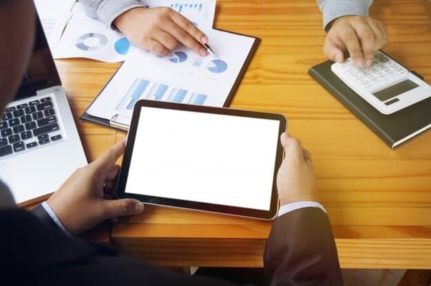 Two people sitting at a table, discussing financial documents with a digital tablet showing investment portfolios in the background.