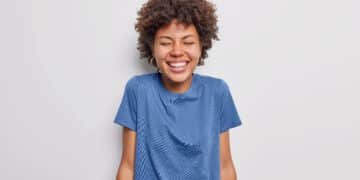 Waist up shot of happy curly haired young woman giggles positively smiles toothily keeps eyes shut wears casual blue t shirt isolated over white background. People and authentic emotions concept