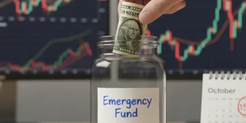 Hand putting money into an emergency savings jar against a background of financial charts.